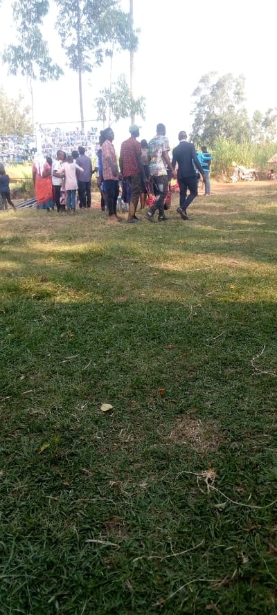 This media file captures an outdoor community gathering in Burundu, Kenya, under bright, clear daylight conditions.
**Subjects**: The primary subjects are a group of approximately 15-20 individuals, mostly adults, with at least one child visible, standing on a grassy field. They are predominantly seen from behind or the side, looking towards a large display board on the left side of the frame. Clothing appears casual for most, with one individual dressed in a dark suit. One woman wears a red dress and a white head covering.
**Objects**: A prominent white board or structure, covered with numerous small photographs arranged in a grid, serves as a focal point for the gathering. The foreground is dominated by lush green grass with some scattered debris. In the background, tall, slender trees with sparse foliage stand against a very bright sky. On the far right, dense green vegetation and what appears to be a dirt path or bare ground are visible, along with a glimpse of a motorbike.
**Scene and Setting**: The scene is set in a sunny, open grassy area, possibly a field or a compound, in Burundu, Kenya. The bright sky suggests a clear day, possibly mid-morning or afternoon, given the shadows on the grass. The tall trees suggest a rural or semi-rural environment.
**Activity or Event**: The people are gathered around and seemingly viewing the display board covered with photographs. This suggests a community event, possibly a memorial, a school function, a local exhibition, or a celebration where images are being showcased or remembered. The general atmosphere appears calm and observant.
**Notable Details**: The intense brightness of the sky indicates strong sunlight. The display board with multiple small pictures is a significant detail, hinting at the purpose of the gathering. There is no clearly visible text on the board or elsewhere that can be deciphered from this distance.