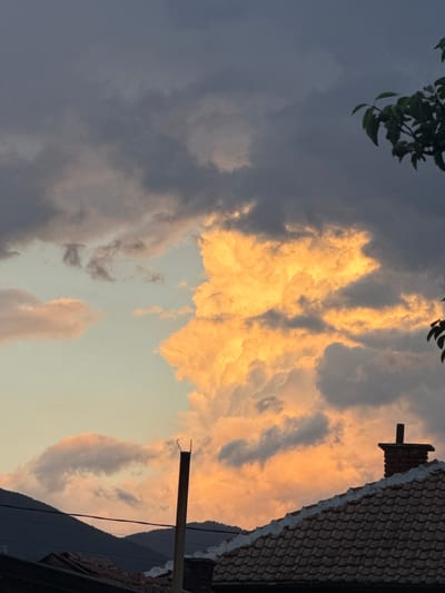 The image depicts a dramatic sky over the city of Kran, Bulgaria. The sky is filled with large, volumetric clouds, some of which are illuminated by a warm, golden-orange light, suggesting sunset or sunrise. Other clouds are dark gray, creating a contrast. In the foreground, the tiled roof of a building is visible, with a brick chimney on the right side. A dark, thin pole, possibly a pipe or antenna, stands vertically in the middle ground. In the background, the silhouettes of dark mountains are visible against the lighter part of the sky. A few dark leaves from a tree are visible in the upper right corner. The overall impression is one of a dynamic and colorful sky over a residential area.