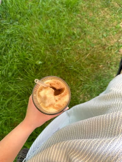 A person's arm and hand hold a glass of a foamy, brown beverage with a clear straw. The glass has a textured surface. The person is wearing a light blue and white striped garment. The background is a blurry green grassy area. The scene is outdoors, with natural light. The location is not specified as Leeds, United Kingdom.