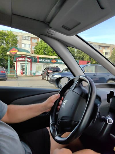 The image captures an interior view from the driver's seat of a car, looking outwards. The driver's hands are on the steering wheel, suggesting they are operating the vehicle. Outside, a small retail building with green awnings is visible, identified by signage as "АЗЕРЖИНСКАЯ" and "МАГАЗИН". A child in a pink outfit stands near the entrance of the building. Several vehicles, including a blue sedan, a black SUV, and a dark blue car, are parked or in motion on a paved area in front of the building. The background shows residential buildings and trees, indicative of an urban or suburban setting. The sky is clear and blue. The presence of an "AIRBAG" label on the car's interior further contextualizes the scene as being inside a vehicle. The location is identified as Fanipal, Belarus.