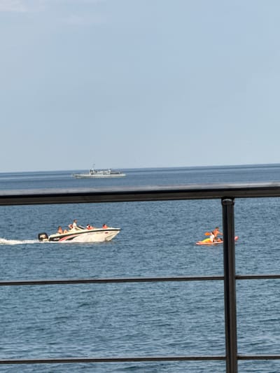 In Partenit, Ukraine, the image captures a serene view of the sea. In the foreground, a metal railing with vertical supports divides the frame, looking out over the blue water. In the middle ground, a white motorboat with people on board is speeding across the water, leaving a white wake. Further in the distance, a person is paddleboarding on a brightly colored board. On the horizon, a larger white ship is visible. The sky above is a clear, light blue.