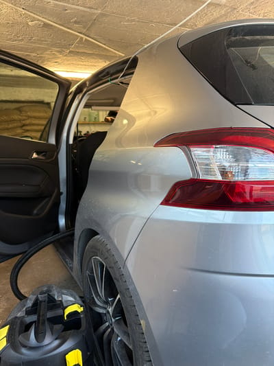 In a garage in Karlovo, Bulgaria, a silver car is being cleaned. The driver's side door is open, revealing the interior. A black vacuum cleaner with yellow accents is positioned on the floor, its hose connected to the car's interior. The car is a hatchback, and the rear taillight is visible. The garage has a textured ceiling, and various items are stored on shelves in the background. The lighting suggests it is daytime, possibly in the evening due to the artificial lights on.