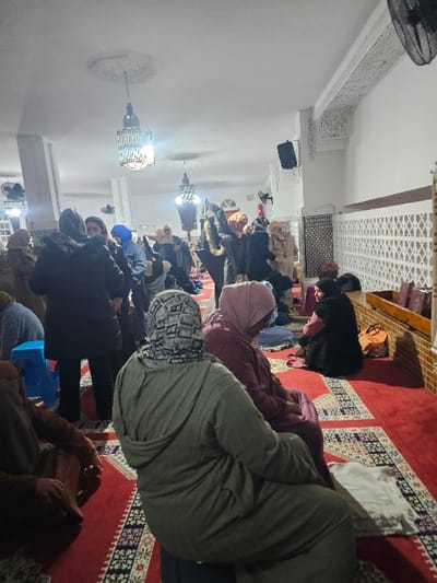 Inside a mosque, a group of people are praying on a red carpet with intricate patterns. Individuals of varying ages and dressed in different attire are kneeling in rows. Some wear head coverings. The mosque has white walls, ornate decorations, and hanging chandeliers. A fan is visible in the upper left corner.
