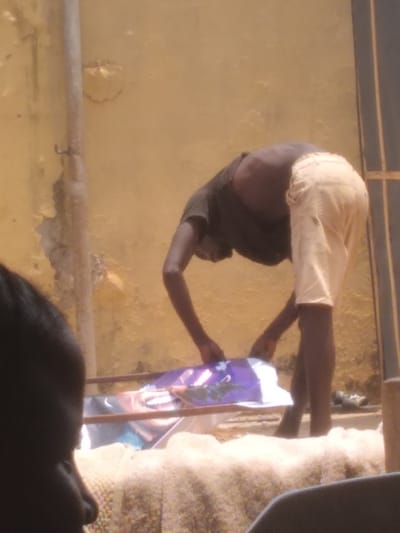 The image portrays an outdoor scene in Agege, Nigeria. A dark-skinned adult male, clad in a dark short-sleeved shirt and light-colored shorts, is bent forward at the waist. His hands are actively engaged with a rectangular object, possibly a banner or poster, featuring a prominent purple design and a printed human face, which rests on a metallic-framed structure. The background comprises a weathered, light yellow wall exhibiting significant peeling paint, with a vertical pipe affixed to its left side. A dark, vertical frame, likely a doorway, is partially visible on the far right. In the blurred foreground, the back of a dark-skinned head and shoulder of a second individual are visible in the bottom left corner. The scene is illuminated by strong natural light.