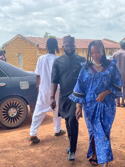 In an outdoor setting with a cloudy sky, three individuals are walking on a dirt path next to a dark blue sedan. A man in the foreground on the right wears a black traditional outfit and a cap, with eyeglasses. Next to him, a woman in a vibrant blue and white patterned dress with ruffled sleeves and a high neckline is walking, adorned with a statement necklace and bracelets. To the left of the man in black, another person dressed in a white traditional outfit is walking away from the camera. The car, identified by a "HONDA ACCORD" emblem, is parked close to the walking individuals. In the background, a single-story building with a red corrugated roof and mud-brick walls is visible. The ground is covered in reddish-brown dirt with scattered pebbles.