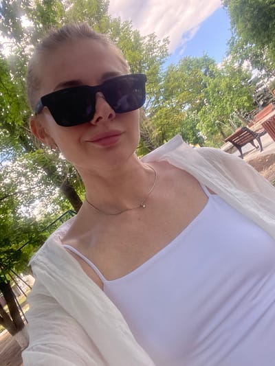 In Markivka, Ukraine, a young woman with blonde hair pulled back and a shaved undercut is captured in a selfie. She is wearing large black sunglasses, a white camisole with thin straps, and an open, lightweight white shirt. A delicate silver necklace with a small pendant rests on her collarbone. The background is a sun-dappled park with lush green trees and a bright blue sky with scattered clouds. Wooden benches are visible in the distance, suggesting a public outdoor space. The lighting indicates it is daytime, likely a sunny afternoon.