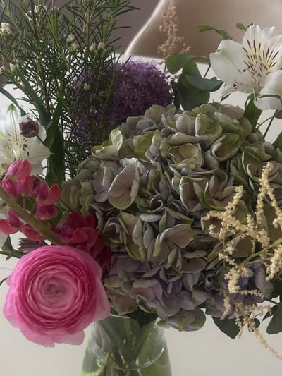 The image shows a close-up of a bouquet of flowers. The bouquet includes a large, round cluster of green and gray hydrangeas in the center. To the left is a purple allium flower. To the bottom right is a pink ranunculus. Other visible flowers are white alstroemeria, small pink lupines and thin green stems with small white flowers. The arrangement appears to be indoors and in a vase. The location is not identifiable.