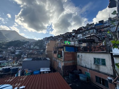 The image displays a panoramic view of a densely populated hillside favela in Rio de Janeiro, Brazil. The sky is partially overcast with large grey and white cumulus clouds, revealing patches of blue sky and bright sunlight shining from the upper left, partially obscured by clouds. In the background to the left, lush green mountains rise, topped by a slender communication tower.
The favela is characterized by numerous multi-story buildings constructed from a mix of raw exposed red brick, light-colored stucco, and weathered concrete. These structures are built closely together, cascading down the steep slope. Many rooftops are flat concrete surfaces, serving as platforms for multiple large, blue, cylindrical water storage tanks and clotheslines with drying laundry. Other roofs are made of corrugated metal, some showing signs of rust.
Two Brazilian flags are visible on a prominent brick building: one larger, horizontally displayed flag featuring green, yellow, and blue, and a smaller one vertically positioned below it. Multiple satellite dishes are affixed to various buildings. On a lower balcony or rooftop, two human figures are discernible. Green vegetation is present in the immediate foreground on the right, framing the scene, and covers the distant mountains.