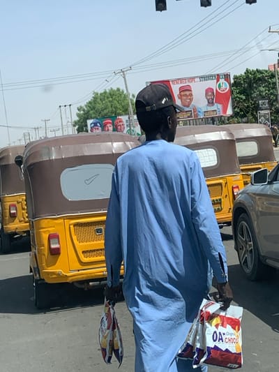 A street scene in Hotoro South, Nigeria, depicts a daytime environment under a clear sky. In the foreground, a dark-skinned male individual, seen from behind, walks right-to-left across a paved road. He wears a light blue long-sleeved tunic and a black baseball cap with a white emblem on the back. He carries a white plastic bag in his right hand, displaying "OA" and "CHOCO" branding, and a multi-colored plastic bag in his left hand. Directly behind him, a line of yellow motorized rickshaws, commonly known as keke napeps, are stopped or moving slowly in traffic. The nearest keke features a brown canopy and a white rectangular marking with a black Nike swoosh on its rear window. A silver vehicle's front fender and wheel are visible on the far right. In the background, power lines traverse the sky, and green foliage is visible. A large billboard displays political messaging with "RENEWED HOPE... STRONGER KA" text and images of four smiling male politicians, primarily in traditional attire. A smaller billboard with additional faces is visible below it. Overhead traffic signals are present.