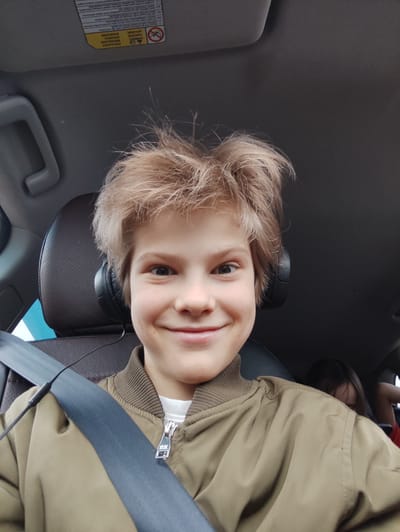 A close-up, eye-level photograph shows a young boy with fair hair wearing a khaki jacket and seatbelt inside a vehicle. He is smiling at the camera. Black headphones are partially visible around his ears. In the background, the headrest of the seat is visible. Behind him, another person is partially visible. The vehicle's interior ceiling is visible in the upper portion of the frame, with a sticker indicating safety information. There is no evidence in the image to place it in the city of Perevoznoye, Russia.