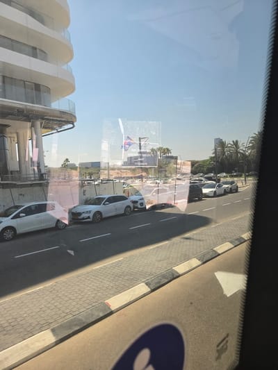 The image depicts a street scene viewed through a window, with reflections visible on the glass. In the foreground, the curved architecture of a modern building is partially visible on the left. The main subject of the image is a street lined with parked cars, predominantly white sedans. These cars are parked along the right side of the road, which is paved with asphalt and bordered by a tiled sidewalk.
In the background, there are palm trees and other greenery, suggesting a warm climate. The sky is clear and blue, indicating a sunny day. The time of day appears to be late morning or early afternoon, given the strong shadows cast by the cars and the sunlight. There are reflections on the window, including what appears to be a person's face and part of another reflection of the interior of a vehicle.
A sign with some text is visible, but it is distorted by the reflection and the angle. Some of the text is in Hebrew. One of the reflections shows a small flag. There is also a large blue circle with a white symbol on the road, likely a road marking or sign. The overall scene suggests a typical urban or suburban environment in a location with a warm climate. The image is taken from inside a vehicle, looking out. The orientation of the image is rotated 90 degrees counterclockwise.