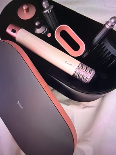 The image shows a close-up, overhead view of a Dyson hair styling kit. The kit is presented in a black, oval-shaped case, with a light pink interior. Several attachments are visible within the case, including what appears to be a main styling wand, brushes, and curling barrels. The styling wand is a light beige color with a rose gold accent at the top and the Dyson logo is printed on it in white. One of the attachments is an oval-shaped ring in a rose gold color. The case itself has a dark gray exterior with a rose gold trim. The kit is placed on a white, textured surface, likely fabric, which is softly creased. There are no people or active events depicted in the image, and no specific location in Beșalma, Moldova, is identifiable.