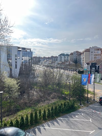 This elevated photograph captures an urban scene along Bulevar Nemanjića in Niš, Serbia, on a bright, partly cloudy day.
In the immediate foreground, the top of a dark grey vehicle, specifically its hood and windshield reflecting sunlight, is visible at the bottom left. Adjacent to this is a paved parking area, featuring light-colored interlocking pavers and white painted parking lines. A red and white striped barrier arm, typical for controlled vehicle access, is positioned at the bottom right. A row of approximately eight small, conical evergreen trees borders the parking lot, casting distinct shadows.
Beyond the parking area, a natural strip of land with varying ground cover, including green grass, bare earth, and scattered sparse vegetation, separates the foreground from the main road. Two dark-colored lampposts are present within this strip.
The midground features a multi-lane asphalt road, Bulevar Nemanjića, with vehicular traffic and numerous parked cars lining both sides. A wide median strip with bare deciduous trees and some early spring foliage divides the road. On the far side of the road, a series of buildings are visible. To the left, a prominent modern building with a distinctive white, angular facade and extensive dark glass windows, identified as "Poslovni Centar Kopernikus," reflects the sky. A blue sign or reflection on its glass appears to show "€ 60." Further right, several multi-story residential and commercial buildings line the street. One ground-floor commercial unit displays a sign reading "PSIHOP CENTAR" (Psychological Center). In front of these buildings, four flagpoles display flags: the leftmost flag is blue with a yellow, red, and white crest; the second is the national flag of Serbia (red, blue, and white horizontal stripes with the greater coat of arms); the third also features red, blue, and white horizontal stripes with an indistinct emblem; and the fourth is a green flag with white text that reads "HELM."
The background consists of additional urban residential buildings, generally light-colored with reddish-brown tiled roofs, extending into the distance under a bright sky with scattered white clouds. No discernible people or specific actions are visible, indicating a static urban landscape.