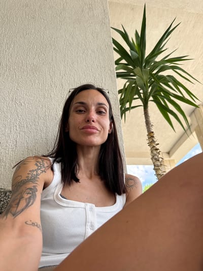 A woman is pictured seated outdoors, leaning against a textured white wall, with a palm tree behind her. She is looking towards the camera with a neutral expression. Her dark hair is shoulder-length, and she is wearing a white ribbed tank top. Tattoos are visible on her left arm and shoulder, including a sun and moon design. The environment suggests a warm climate.
