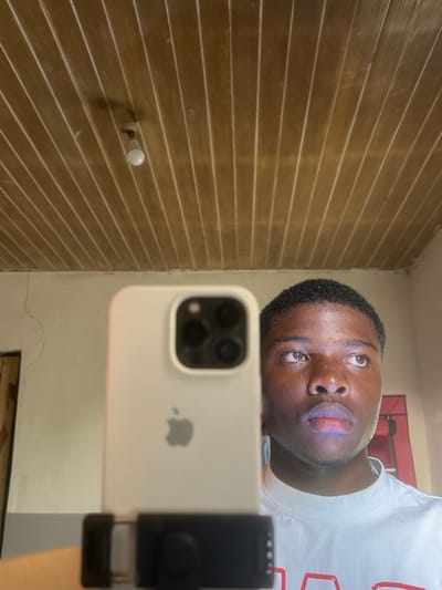 The image depicts a young male taking a selfie in what appears to be an indoor room in Bar Gada, Nigeria. The subject, a male with dark skin and short, dark, closely cropped hair, is positioned in the right half of the frame, looking slightly upward and to the right. His face is illuminated from the front, with a distinct light reflection visible on his forehead and lips. He is wearing a white t-shirt. In the left half of the frame, a light-colored iPhone, likely an iPhone 14 Pro or 15 Pro, encased in a light-colored protective cover, is held vertically. The Apple logo and the three-lens camera array are visible. The phone is secured in a black clip-style tripod mount, which is partially visible at the bottom. The background features a paneled wooden ceiling with light brown planks running longitudinally. A single white LED light bulb hangs from the ceiling, slightly left of center. The walls are light-colored, possibly off-white, with visible cracks on the left wall. A dark opening, potentially a doorway, is partially visible on the far left. A red-framed object is faintly visible on the far right.