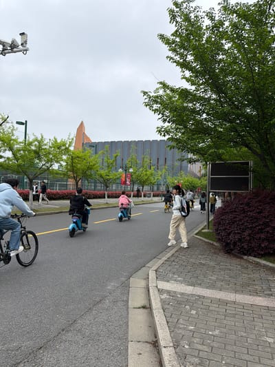 The image shows a street scene in the Jiangning District of China, likely on a university campus. The sky is overcast. A paved road runs through the scene with a yellow line indicating a lane. Several people are on the road, including one person riding a bicycle in the foreground. Further up the road, several people are riding small blue electric scooters. Two individuals are walking alongside the road on a paved area. Buildings with modern architecture are visible along the side of the road, with greenery including trees and bushes. A signpost is next to the paved walkway.