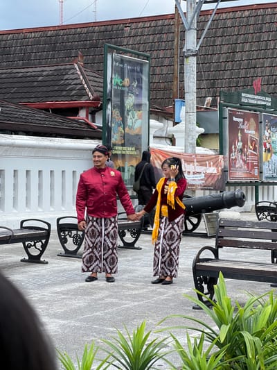 A man and woman in traditional Indonesian attire pose beside a bench and cannon. The man wears a red jacket and a traditional headpiece, while the woman sports a reddish-brown top and a yellow sash. Both wear similar patterned batik sarongs. Informational posters line the wall in the background, and plants appear on the right.
