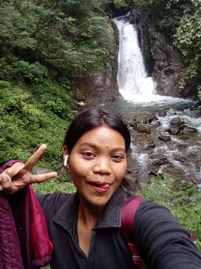 The image shows a young woman standing in front of a waterfall in the Grojogan Sewu waterfall area, located in the Tawangmangu District of Karanganyar Regency in Central Java, Indonesia. The woman has dark hair and is wearing a black jacket with a red stripe on the strap of her backpack. She is holding up two fingers in a peace sign gesture and is smiling at the camera. Her tongue is sticking out. The waterfall is in the background, cascading down a rocky cliff face. The area around the waterfall is lush and green with trees and foliage. There is also a stream of water flowing in the foreground, along with rocks and vegetation. The coordinates indicate a location near the base of the waterfall.  The image is a selfie taken from a close-up perspective.  It is likely taken by the woman herself. The overall impression is one of a pleasant, carefree moment in nature.