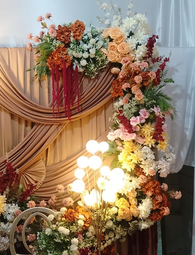 The image depicts a decorative floral arrangement at what appears to be an event venue in Cibrek, Indonesia. The arrangement is elaborate and features a variety of flowers in shades of peach, cream, white, pink, and rust orange, interspersed with greenery. Prominent elements include cascading strings of deep red flowers that hang like tassels, and large, bulbous white lights arranged on a stand in the foreground.

The background consists of draped fabric, with a golden-brown material forming a pleated backdrop on the left and white sheer curtains on the right. The lighting is warm and suggests an indoor setting, possibly in the evening or with artificial lighting designed to create a cozy ambiance. The overall impression is one of festivity and decoration, likely for a wedding, party, or other celebratory occasion. There are no people visible in the image, and no text is present. The focus is entirely on the floral display and the surrounding decor.