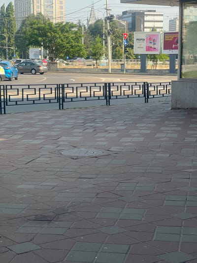 The image shows a street scene in Iasi, Romania, with a view of a sidewalk, a street with cars, and buildings in the background. A black metal fence with a geometric pattern runs horizontally across the middle of the image, separating the sidewalk from the street. The sidewalk is paved with interlocking geometric tiles in shades of red and grey, some of which are uneven or broken. A manhole cover is visible on the sidewalk.
In the street, several cars are visible, including a blue car and a grey sedan. There is a traffic light and a pedestrian crossing. In the background, there are trees and several buildings, including a modern office building with a glass facade and another building with a classical architectural style. Billboards are also visible, one of which advertises Pepsi. The sky is clear and appears to be daytime. The overall impression is an urban environment on a sunny day.