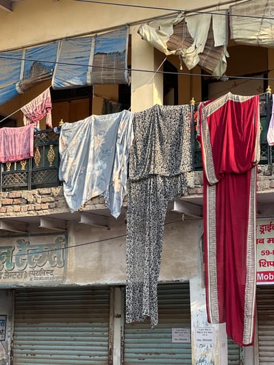 The image depicts an outdoor urban scene in Mathura, India, characterized by the presence of various textiles hung on a wire between buildings. At the top, a red garment with intricate borders is visible, followed by a black and white floral patterned fabric, and a blue floral patterned sheet hanging below. These items appear to be drying or displayed outdoors.
The buildings framing the scene showcase weathered facades. One has metal shutters and signs with Hindi script, including the name “निरन्जन लाल अर्जुन पुरा मधुर” and the phrase “ये दुकान चौराह पर शिफ्ट हो गई है परिक्रमा मार्ग पर" along with a mobile number (7983316491).
Above the textiles are makeshift awnings or shades made of tattered fabric and bamboo, attached to the buildings. The environment appears cluttered with wires running across the scene and aged brickwork.