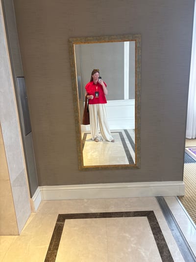 In the reflection of a rectangular, ornate mirror, a person is seen standing. They are wearing a bright red top and loose, cream-colored pants. A brown bag is slung over their shoulder. The person appears to be holding a phone in their hand, likely taking the picture. They are also wearing sunglasses.

The mirror hangs on a textured, taupe-colored wall. Behind the person, the reflection shows a hallway with a white door and a floor composed of light-colored tiles with dark gray inlaid geometric patterns. The overall lighting suggests it is daytime. The setting appears to be indoors, possibly a hotel lobby or a well-decorated corridor in a public building. There is a white trim running along the wall and floor, separating different interior finishes. Visible text on a lanyard or badge around the person's neck includes a QR code. The image is rotated 90 degrees clockwise.