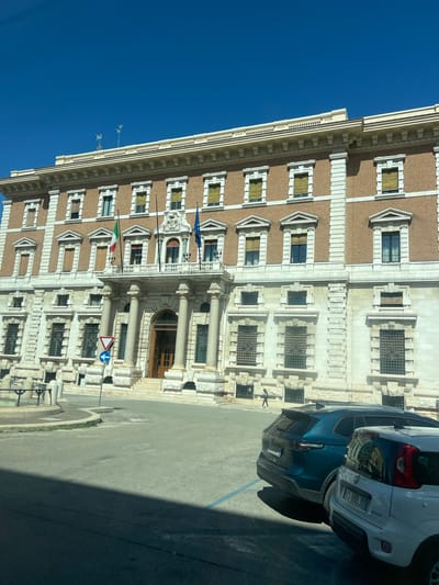The image shows the Banca d'Italia building in Bari, Italy, on a clear, sunny day. The facade of the building is made of brick and stone, with multiple rows of windows. Two flags, the Italian flag and the European Union flag, are flying from the balcony above the main entrance. Several cars are parked in the foreground, and a person is walking on the sidewalk in front of the building. The street in front of the building is paved, and there is a traffic sign on the left side of the image.