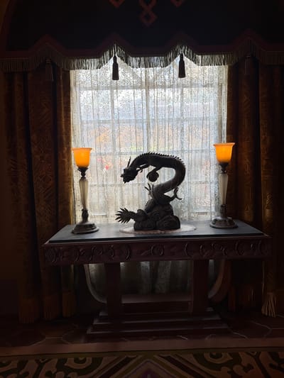 The image depicts an interior scene, likely in Chessy, France, featuring a window with ornate curtains and a carved wooden table. A dragon sculpture is prominently displayed on the table, positioned in front of the window, which is covered by a sheer, patterned fabric. Two lamps with orange glass shades are mounted on the table, one on each side of the dragon sculpture. The curtains are heavy and dark, with decorative fringe and tassels, framing the window. The overall aesthetic suggests a dramatic or theatrical setting, possibly a hotel lobby or a themed room. The lighting is dim, with the lamps providing a warm, ambient glow, and the sunlight filtering through the window creates a contrast between the illuminated background and the darker foreground. There is no visible text in the image.