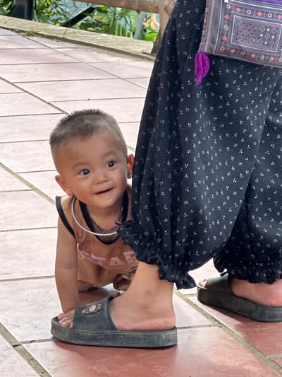 In Tu Le, Vietnam, a young child, approximately 1-2 years old, is captured in a crawling or crouching position, peeking from behind an adult's left leg. The child has short, dark hair, tan skin, and wears a sleeveless, light brown top with a dark trim. A thin, silvery-white metal ring rests around the child's neck. The child looks directly at the viewer with a curious, open-mouthed smile. The adult's lower legs are clad in dark, patterned trousers or a long skirt featuring small, light-colored repeating motifs, with ruffled hems above the ankles. The adult wears dark, flat slide sandals; the left sandal has a small, light-colored emblem. A patterned, woven bag with intricate geometric designs in red, purple, black, white, and yellow/gold, accented by a purple tassel, hangs from the adult's hip. The subjects are standing on a reddish-brown tiled surface with lighter grout lines. In the blurred background, green foliage and a light-colored concrete ledge are visible, suggesting an outdoor setting.