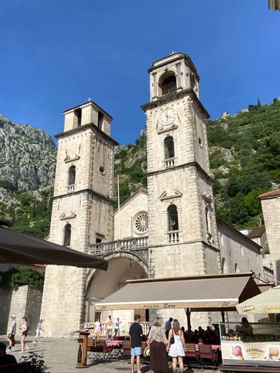 The image displays the Cathedral of Saint Tryphon in Kotor, Montenegro, under a clear blue sky, with rugged, green-covered mountains rising immediately behind it. The cathedral is constructed from light-colored stone and features two prominent bell towers. The right tower contains a circular clock face indicating approximately 12:50 and an arched belfry opening at its peak. The left tower also has an arched belfry opening and is topped with a small white cross. A large circular rose window is centered on the cathedral's facade between the two towers, above a substantial arched entrance. A stone balustrade connects the two towers across the facade. The year "2016" is carved into the stone base of the right tower.

In the foreground, a paved public square is active with pedestrians and outdoor dining. A large, retractable brown awning from "Pizzeria Sara" extends over multiple tables and chairs, under which several people are seated or standing. To the right, an outdoor ice cream stand with a glass display freezer is visible, featuring "PREMIUM ICE CREAM" and "Frikon" branding. Numerous individuals are traversing the square; one person in a white dress and another in a long brown skirt are walking towards the "Pizzeria Sara" area. Other people are dispersed across the square, engaged in walking or standing. A white flagpole stands between the cathedral towers.