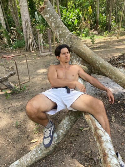 In Los Velásquez, Venezuela, a shirtless man sits casually on a fallen tree trunk in a wooded area. He is wearing white shorts and athletic shoes. The man is looking off to the side with a pensive expression. The background consists of lush green foliage and more trees, suggesting a natural, outdoor setting. The ground is covered in dirt and fallen leaves. There is a rusty metal structure to the left of the man.