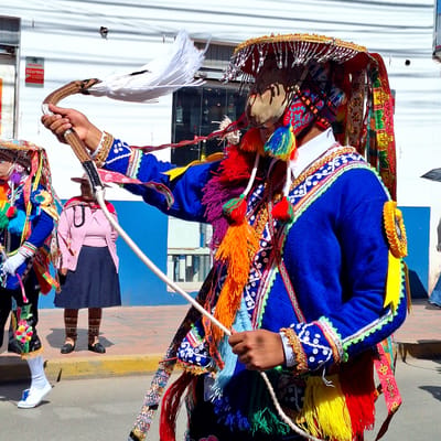 The image shows a person in traditional Wanchaq, Peru attire, likely participating in a festival or cultural event. The subject is adorned in a vibrant blue tunic with ornate, colorful embroidery featuring intricate patterns and gold trim. The costume includes a wide, richly decorated hat with fringes and tassels in yellow, red, and blue. The person is holding a whip-like object with a white feathery end in their right hand, and a white rope in their left hand, suggesting they are performing a dance or ritual.

The background features a white corrugated wall on the left, and a textured paved ground with a yellow curb on the right. Sunlight suggests it is daytime, likely under clear weather conditions. Reflections on the ground indicate a sunny day. The overall scene is lively, implied by the colorful costume and energetic posture. The person's face is partially obscured by the mask and headdress, but the overall impression is one of focused participation.

Below the main subject, another individual in a similarly colorful, albeit less elaborate, costume is visible, suggesting a group performance or procession. There are some shadows cast on the ground, indicating the direction of the sun. A red and white "Alarma" sticker with the Verisure logo is visible on the white wall, indicating a nearby commercial establishment. The context of Wanchaq, Peru, strongly suggests a folk celebration, possibly a religious or agricultural festival.