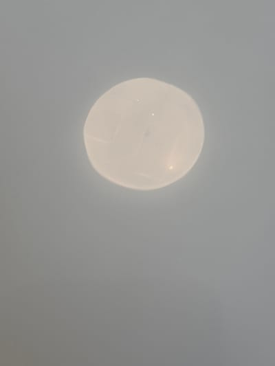 A close-up, out-of-focus image shows a round, translucent object with faint internal markings resembling the panels of a volleyball. The object is brightly lit from within or by an external light source, causing it to appear luminous. The background is a uniform, soft grey. There is no discernible context or location in the city of Zagrađe, Montenegro.