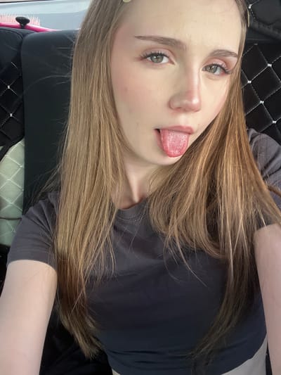 A young woman with long, light brown hair is pictured from the chest up, taking a selfie. She is wearing a dark grey t-shirt. Her right eye is looking directly at the camera, while her left eye is looking slightly off to the side. Her tongue is sticking out. She has light makeup on, including pink blush and mascara. The background appears to be the interior of a vehicle, with a black quilted seat visible to the right of the woman and a lighter patterned seat to the left. There is a pink hairbrush with a black handle on the left side of the frame, resting on the black seat. The location is identified as Novaya Otradovka, Russia.