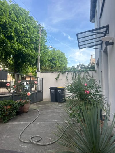 The image displays a bright, sunny outdoor courtyard or patio area, viewed from a slightly elevated position. On the right side, a white building wall features a dark metal awning with translucent panels and a white circular outdoor light fixture. Below this, a large palm-like plant with prominent green fronds is a central feature, near which a rose bush with several pink blooms adds a touch of color. Two dark grey/black garbage bins with yellow lids, distinctly labeled "Paris Est", are placed against the wall.
The concrete ground covers the bottom and mid-sections of the scene, with a grey garden hose coiled at the bottom right and extending across the patio. Along the lower edge, several potted plants, including another rose bush with pink flowers and some ivy, are visible. A dark metal gate or fence spans the middle ground, marking a boundary to a dense expanse of lush green trees that dominate the left side of the image.
A utility pole with overhead power lines is visible amidst the trees on the far left. The background is a clear blue sky with scattered white clouds and faint contrails. The white wall further back is partially covered with climbing greenery. The scene is bathed in bright daylight, suggesting a clear day. The text "Paris Est" on the bins indicates a location in the eastern part of Paris, aligning with Joinville-le-Pont, France. No people or explicit activities are depicted.
