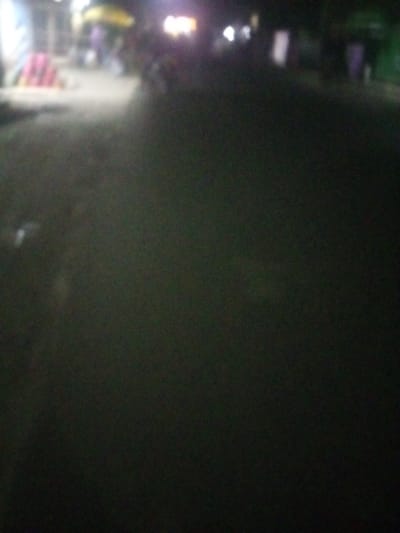 The image captures a poorly lit outdoor scene at night. The foreground is dominated by a dark, textured surface, possibly a road. In the distance, blurred lights and indistinct shapes suggest a building or structure, with hints of activity or parked vehicles. The overall mood is one of darkness and obscurity, making it difficult to discern specific details.