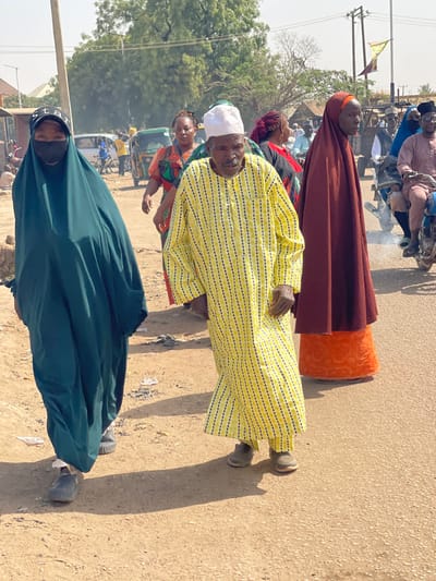 The image depicts an outdoor street scene in Chikaji, Nigeria, under bright daylight. In the foreground, three individuals are walking. On the left, a person wearing a dark teal abaya, a black face mask, and a black baseball cap with white "STAR" lettering walks towards the left, with dark shoes visible. Centrally positioned, an elderly man in a long-sleeved yellow garment with black vertical stripes and a white cap walks towards the right, his right hand slightly raised, wearing light brown shoes. To the right of the elderly man, another person is partially visible, clad in a dark red head covering over an orange patterned dress, also walking towards the right.
In the mid-ground, a woman in a patterned top is visible behind the elderly man, looking left. Further back, a green auto-rickshaw is parked on the left side of the dirt road, while multiple individuals, some riding motorcycles or scooters emitting exhaust smoke, are present on the right side of the road. The environment includes a dry, light brown dirt road surface, utility poles with wires, sparse trees, and low-rise buildings or structures in the background. Strong sunlight illuminates the scene, casting distinct shadows.