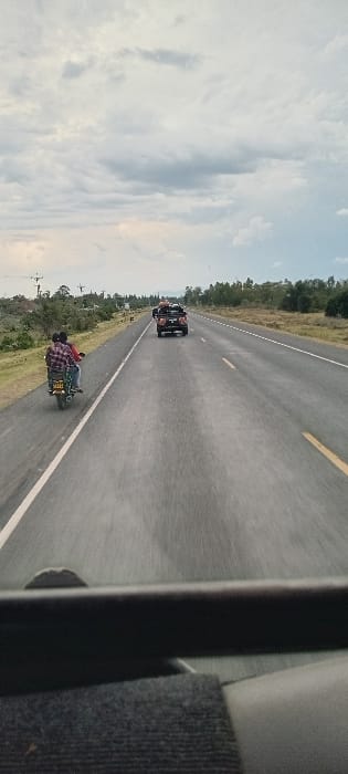 An open highway is pictured, with a vehicle visible in the distance and a motorbike riding to the side of the road. The sky is slightly overcast with a few clouds, and the edges of the road are bordered by foliage. The photo is taken from inside a vehicle, with part of the interior visible in the right frame.
