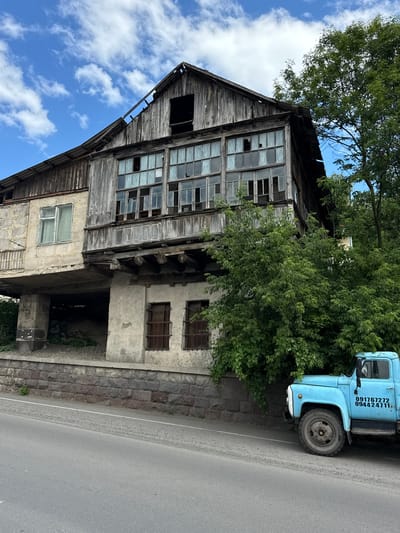 In the city of Dilijan, Armenia, a partially dilapidated wooden and concrete building stands beside a road. The building has multiple stories, with the upper levels featuring wooden siding and numerous broken windows. A lower section of the building is made of concrete and appears more weathered. A lush green tree grows in front of the building, partially obscuring it. To the right of the building, a bright blue pickup truck is parked on the side of the road, facing towards the right. The truck has two phone numbers printed on its side. The road is paved with asphalt, and a stone wall runs along the base of the building. The sky above is blue with scattered white clouds.