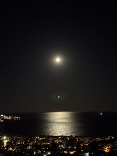 An aerial photograph captures the night scene in Šušanj, Montenegro. The full moon dominates the sky, with its light reflected on the still water below. The city lights from the buildings along the shore illuminate the coastline. The photograph does not show people or specific actions; instead, it captures the overall serene atmosphere of a coastal city at night.