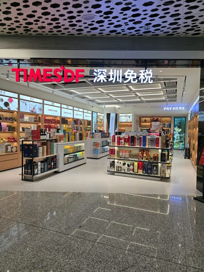 The image shows the interior of a duty-free shop in Bao'an District, China. The shop is well-lit with a modern design. Above the entrance, a prominent red sign reads "TIMES DF" followed by Chinese characters that translate to "Shenzhen Duty Free." Inside, shelves are stocked with various products, including liquor, cosmetics, and gift boxes. Several display shelves are visible, showcasing bottles of wine and spirits, as well as packaged goods. A digital screen displaying "PAY HERE" is mounted on the wall. The floor is made of polished granite tiles with inlaid lighting. No people are visible in the image. The overall environment suggests a retail setting, likely an airport or a shopping mall.