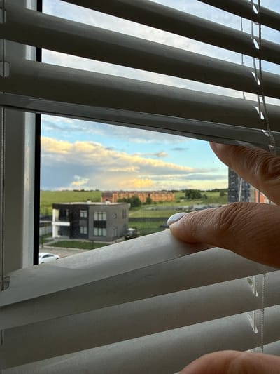 The image shows a view from a window through partially open white blinds. A person's hands are visible, adjusting the blinds. The scene outside depicts a suburban or semi-urban landscape with modern buildings, green spaces, and a cloudy sky with hints of sunset colors. The architecture suggests a contemporary development, potentially in a region like Vologda, Russia, where such construction styles are common. The overall impression is one of looking out from a dwelling, with a focus on the act of partially revealing the outside world.