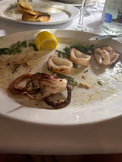 The image shows a white plate with remnants of a meal, including pieces of grilled squid and a slice of lemon. The plate is partially obscured by a tablecloth. In the background, another plate with toast can be seen. The scene appears to be from a restaurant in the Sithonia Municipal Unit of Greece, indicated by the presence of Greek food and tableware.
