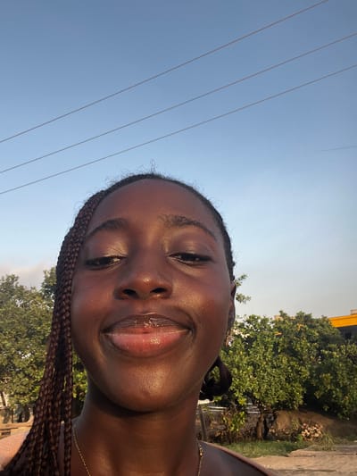 A young woman with dark skin and braided hair is taking a selfie outdoors. She is smiling with her lips slightly parted, revealing lip gloss. In the background, there are green trees, power lines against a blue sky, and a yellow building. The image appears to be taken in Karum, Nigeria.