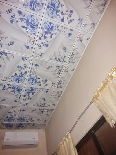 The image shows a wall covered with square decorative tiles featuring a blue floral pattern on a white background. These tiles appear to be positioned in a grid-like arrangement. Adjacent to the tiled wall is a small, white, rectangular object, likely an air conditioning unit or similar appliance. The ceiling is visible above the tiled wall and features a decorative element, possibly a curtain or some sort of fabric draping. The environment appears to be indoors, with the lighting suggesting an enclosed space. The image's angle offers a perspective from below, looking upwards at the tiled wall and ceiling. There are no people visible in the frame. Without further information, the location in Santa Cruz de la Sierra cannot be determined.