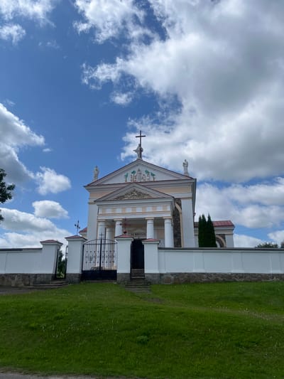 The image depicts the facade of the Church of the Transfiguration in Zhaludok, Belarus, under a partly cloudy blue sky. The church is a Neoclassical building with a prominent pediment topped with a cross and statues. The facade features a portico with columns and decorative elements. A white wall surrounds the church grounds, with a black wrought-iron gate in the foreground. A well-maintained green lawn stretches out in front of the wall. The overall scene is one of a serene, historical religious site bathed in daylight.