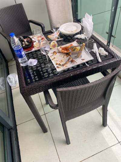 A rectangular dark wicker table with a glass top is heavily laden with the remnants of a meal. A white rectangular plate displays food scraps, including what appears to be fried food remnants and sauces. Several used napkins are present, along with a partially consumed bottle of water and various empty glasses. The scene is outdoors, as indicated by the tiled floor and the presence of reflective glass surfaces in the background. A matching wicker chair is positioned beside the table. There are no people visible in the image. There is no information about the location in the city of Itirin, Nigeria.