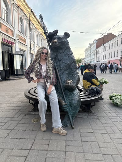 The image captures an urban street scene in Заволжье, Russia, featuring a young woman posing next to a large bear statue. The woman, with long blonde dreadlocks and sunglasses, is dressed in a red and black plaid shirt over a light top, light-wash jeans, and beige sneakers. She is smiling and looking towards the camera, holding a small white pouch.
To her right, a dark-colored statue of a bear sits on a decorative bench, holding a staff. A smaller, lighter-colored animal figure rests on the bear's left leg. In the background, another person wearing a yellow and black jacket is seated on the bench, holding a bouquet of white flowers. Several other pedestrians are visible walking along the grey-tiled sidewalk.
The street is lined with multi-story buildings, displaying varying architectural styles with numerous windows and decorative elements. The sky appears bright and partly cloudy. On one of the buildings in the background, a sign with Cyrillic text is visible, clearly showing "СЛАДОСТИ" (SLADOSTI), meaning "Sweets" or "Confectionery". The scene depicts a casual daytime moment, likely in a pedestrian area of the city.