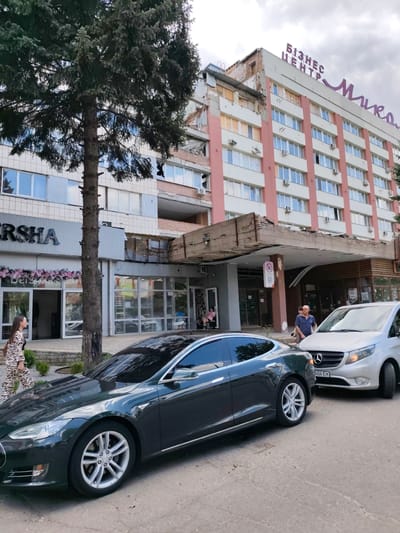 The image depicts a street scene in Mykolaiv, Ukraine. In the foreground, a dark-colored Tesla Model S is parked on the right side of the road. To its right, a silver Mercedes-Benz Vito van is also parked. A man in a blue shirt and sunglasses stands near the van, looking towards the right.

In the middle ground, a woman with a zebra-patterned dress walks on the sidewalk in front of a store with a large "BERSHA" sign. The store has glass doors and displays decorated with pink flowers. Adjacent to this store is another entrance with a sign that appears to be an emblem.

Dominating the background is a large, multi-story building with a sign at the top that reads "БІЗНЕС ЦЕНТР Миколаїв" (Business Center Mykolaiv). The building shows signs of damage, particularly on the upper floors and the overhang above the entrance. Some windows are broken or boarded up, and there is visible debris on the overhang. A large evergreen tree is positioned in the left foreground, partially obscuring the view of the building. The sky is overcast.