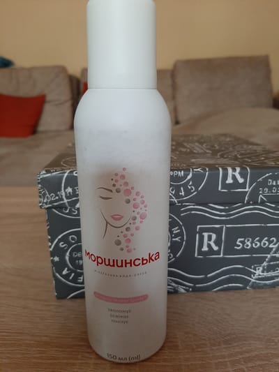 The image shows a white aerosol can with pink and gray circular patterns and a stylized face on it, placed on top of a patterned box. The box has a design resembling postage stamps with the letters "R", "N.Y.", and numbers like "58662". The can appears to be a beauty product, possibly a mineral water spray, as indicated by the text on it. The text on the can is in Ukrainian and English. In Ukrainian, it reads "Моршинська" (Morshynska), "МІНЕРАЛЬНА ВОДА-СПРЕЙ" (Mineral Water Spray), and "зволожує освіжає тонізує" (moisturizes, refreshes, tones). The English text also says "Mineral Water Spray". The volume is stated as "150 мл (ml)". The patterned box has text like "Dat 20.05", "02.19", and "PM". The overall scene is indoors, with a wooden surface visible in the foreground and a blurred background suggesting furniture like a sofa with orange cushions. The lighting appears to be artificial and the setting is casual. The image is oriented vertically, with the can angled slightly upwards.